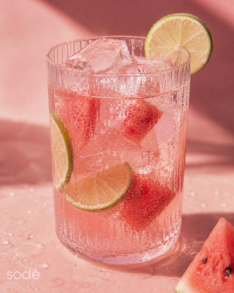 A clear ribbed drinking glass filled with a pink iced beverage, watermelon cubes, lime slices, and ice cubes, set on a pink surface with a matching pink background.